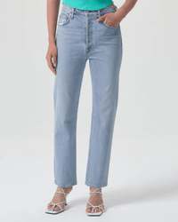 Wearing AGOLDE’s High Rise Stovepipe in Destination jeans, a green top, and white strappy heeled sandals, a person stands with one hand in their pocket against a plain background.