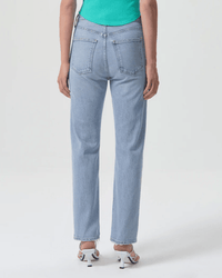 Someone is shown from the back wearing AGOLDE's High Rise Stovepipe in Destination denim, a teal top, and white heeled sandals, standing against a plain light background.