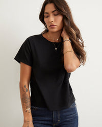 A woman with long wavy brown hair and tattoos on her left forearm stands against a light background, wearing the AMO Classic Tee in Black and dark jeans. She touches her hair with her right hand and looks down thoughtfully.