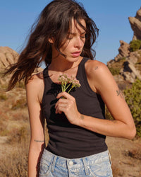 A woman with long brown hair wears the AMO Long Rib Tank in Vintage Black and blue jeans, standing outdoors among rocky desert scenery. She holds a small bouquet of wildflowers and gazes down thoughtfully.