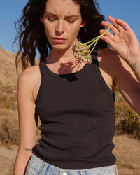 Wearing the AMO Long Rib Tank in Vintage Black and light denim jeans, a woman stands outdoors, holding pink flowers and looking down seriously amid dry grass and rocky hills.