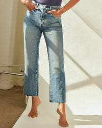 A person stands on tiptoe, wearing AMO's Loverboy in Loved high rise straight leg jeans with a raw hem ankle crop and a tucked-in purple top, set against a softly lit indoor background.