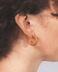 Close-up of a person’s ear wearing the B&L C Hoop in Mini in Leather Weekender: a thick, round, golden-brown mango wood earring. The fair skin and dark wavy hair partially frame the handmade, sustainably sourced piece against a neutral background.