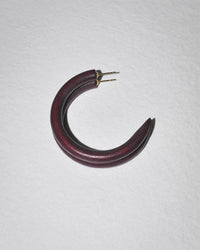 A single B&L C Hoop in Small in Cocoa, a handcrafted dark red crescent-shaped earring with gold posts, rests on a textured white surface.