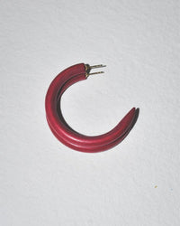 A single B&L C Hoop in Small in Pinto, a red crescent-shaped mango wood earring with a glossy finish and two metal posts, rests on a white textured surface.