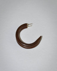 A single Small Hoop in Raisin by B&L, a dark brown crescent-shaped earring made from sustainably sourced mango wood with gold posts, rests on a plain light gray surface.