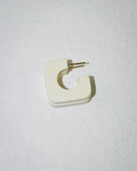 The Square Hoop in Chanel Ivory by B&L is a single, handmade geometric earring with an open square design, crafted from sustainably sourced materials. It features an off-white finish and a gold post, shown resting on a textured white surface.