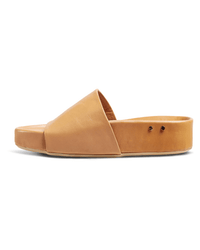 The beek.. Pelican in Honey is a tan leather slide sandal with a wide diagonal strap, open toe, flat sole, and two small metal rivets—designed for comfort and arch support. Shown against a white background.
