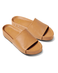 The Pelican in Honey by beek. is a pair of tan leather slide sandals with wide straps, flat soles, and subtle arch support, shown diagonally on a white background.