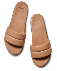 A pair of tan slip-on sandals, the Sugarbird in Beach by beek., with wide padded straps and flat soles, shown from above on a white background. The comfortable leather slides feature an insole stamped “beek.” in cursive.