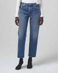 Person wearing Citizens of Humanity Neve Low Slung Relaxed jeans in Oasis, a white ribbed long-sleeve top, black socks, and black shoes, standing against a plain light grey background. Photo is cropped from the shoulders down.