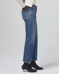 A person wears Citizens of Humanity Neve Low Slung Relaxed jeans in Oasis, a white ribbed sweater, black socks, and black open-toe kitten heels, standing against a plain light background.