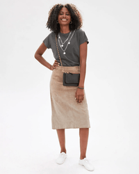 A woman smiles against a white background, wearing a dark gray T-shirt, beige suede midi skirt, white sneakers, layered necklaces, and a Clare V. crossbody purse with the Box Chain Crossbody Strap in Brass.