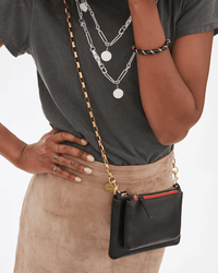 A person in a grey t-shirt, beige suede skirt, layered silver jewelry, and rings holds a Clare V. bag with a Box Chain Crossbody Strap in Brass and a bold red zipper detail.