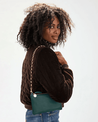 A woman with curly hair in a dark quilted jacket and blue jeans looks over her shoulder, holding a Clare V. purse featuring the Box Chain Shoulder Strap in Brass.