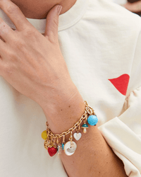 A person in a cream shirt with a red heart detail touches their neck while wearing the Clare V. Charm Chain Bracelet in Vintage Gold, decorated with colorful charms like a strawberry, blue bead, shell, and other small pendants.