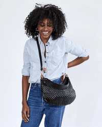 A smiling woman with curly hair models the Clare V. Moyen Messenger in Black Zig Zag, wearing a light blue button-up shirt and blue jeans, holding the bag by its crossbody strap against a plain white background.