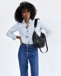 A woman with curly hair wears a light blue shirt, blue jeans, gold jewelry, and carries the Clare V. Moyen Messenger in Black Zig Zag over her shoulder, standing with one hand on her hip against a white background.