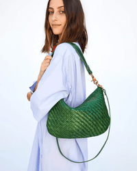 A woman in a light blue button-up shirt holds the Clare V. Moyen Messenger - Woven Zig-Zag in Evergreen, featuring gold hardware, smiling slightly against a plain light background.