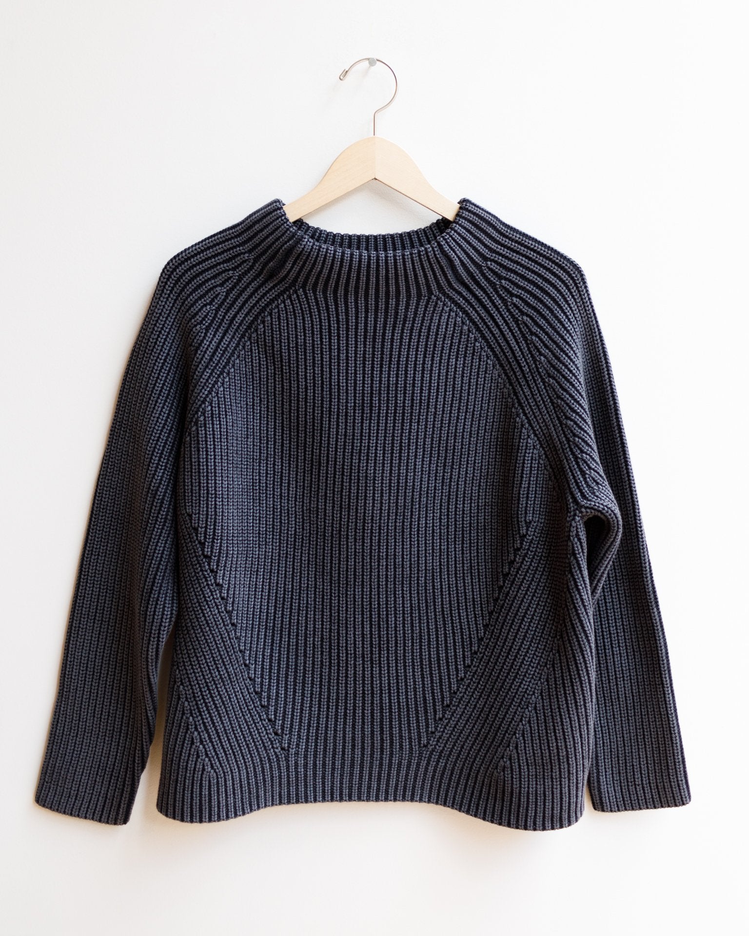 The Demylee Daphne Washed Cotton Sweater in Black, featuring classic Fisherman-inspired ribbed knit and long sleeves, hangs on a wooden hanger against a plain white background.