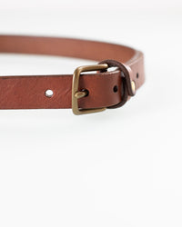 Close-up of the Depalma Handsewn 1 inch Mon Senor Belt in Bark with brass buckle, crafted from genuine leather. The belt lies flat on a white surface, showing adjustment holes and a leather loop near the solid brass buckle.