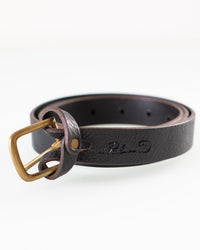 A coiled 1 inch Mon Senor Belt in Black w/ Brass by Depalma Handsewn features a brass buckle, fine stitching, and subtle embossed markings on the strap, all set against a plain white background.