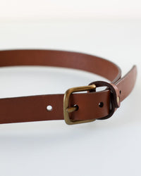 A close-up of the Depalma Handsewn 1 inch Mon Senor Belt in Tobacco with a solid brass buckle, partially coiled on a white surface.