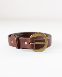 The CL 1 inch Belt in Bark/Brass by Depalma Handsewn, featuring brown genuine leather, a gold oval brass buckle, and two metal rivets, is coiled and displayed on a white surface.