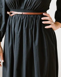 A person wears a black dress styled with the Depalma Handsewn Nouva 1/2 inch Single Wrap belt in Bark/Brass, hand on hip to show off manicured nails and a ring. The photo is cropped at the shoulders and below the waist.