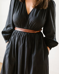 A woman stands with her hands in her pockets, wearing a black long-sleeved dress with a V-neckline, styled with the Depalma Handsewn Nuova 1/2 inch Dbl Wrap belt in Bark/Brass at the waist. Her head is not visible in the frame.