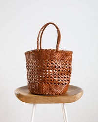The Cannage Bucket Big in Tan by Dragon Diffusion, featuring an open cannage weave and two handles, is displayed on a wooden stool with white legs against a plain white background.