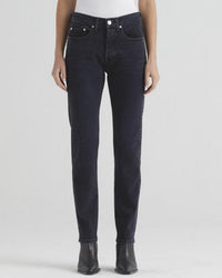 A person wearing Edwin’s Hana High Rise Straight jeans in Goth, paired with a white top and black ankle boots, stands against a light gray background. The look highlights the dark blue high-waisted straight-leg jeans.