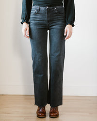A person stands indoors on a wooden floor against a white wall, wearing Edwin's Marli in Fog jeans—high-rise, dark wash with a raw hem—and brown leather shoes. Their upper body is partially visible.