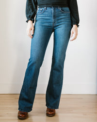 A person wears Edwin’s Ryder in Tangled high rise bootcut jeans (medium indigo vintage wash), a black long-sleeve top, and brown shoes, standing on a light wooden floor against a white wall. Photo is cropped at the shoulders.