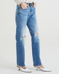 A person wears Edwin’s Tash in Symbol 90s mid-blue relaxed jeans with a belt and white heeled shoes, standing against a plain white background. Only their lower half is visible.