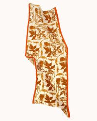 The Épice Botanical Print Scarf in Golden is a rectangular handwoven linen-cotton stole with an off-white base, brown botanical and floral Danish herbarium prints, and a bright orange border, shown slightly folded and draped.