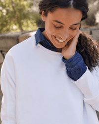 A person with long dark hair, wearing the Frank & Eileen Anna Long Sleeve Fleece Capelet in White over a blue collared shirt, smiles while looking down and touching their face, with greenery and a stone wall blurred in the background.