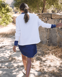 A woman with curly hair tied back walks on a stone path by a rock wall and trees, wearing the Frank & Eileen Anna Long Sleeve Fleece Capelet in White over a blue dress and white sneakers in a sunlit outdoor setting.