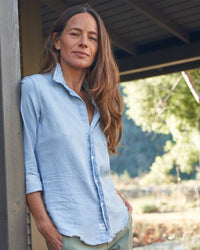 A woman with long brown hair wears the Frank & Eileen Barry Button Up in Classic Blue w/ Tattered Wash as she stands outdoors, leaning against a wooden post with sunlit trees behind her.