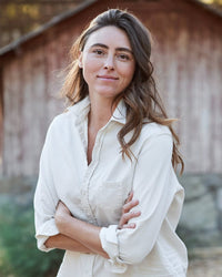 A woman with long brown hair stands outdoors, smiling softly with arms crossed, wearing the Frank & Eileen Eileen Woven Button Up in Vintage White. A rustic wooden building is blurred in the background.