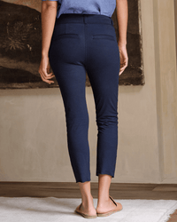 A person stands indoors facing away, wearing Frank & Eileen's Murphy Pant in British Royal Navy styled as leggings with beige flats; arms relaxed by their sides. The background shows a neutral wall and partially visible framed artwork.