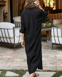 A woman with wavy hair, in the Rory Woven Long Dress in Black Linen by Frank & Eileen and sandals, stands outdoors on a tiled patio with her back to the camera near white cushioned iron chairs and a wooden table with plants.