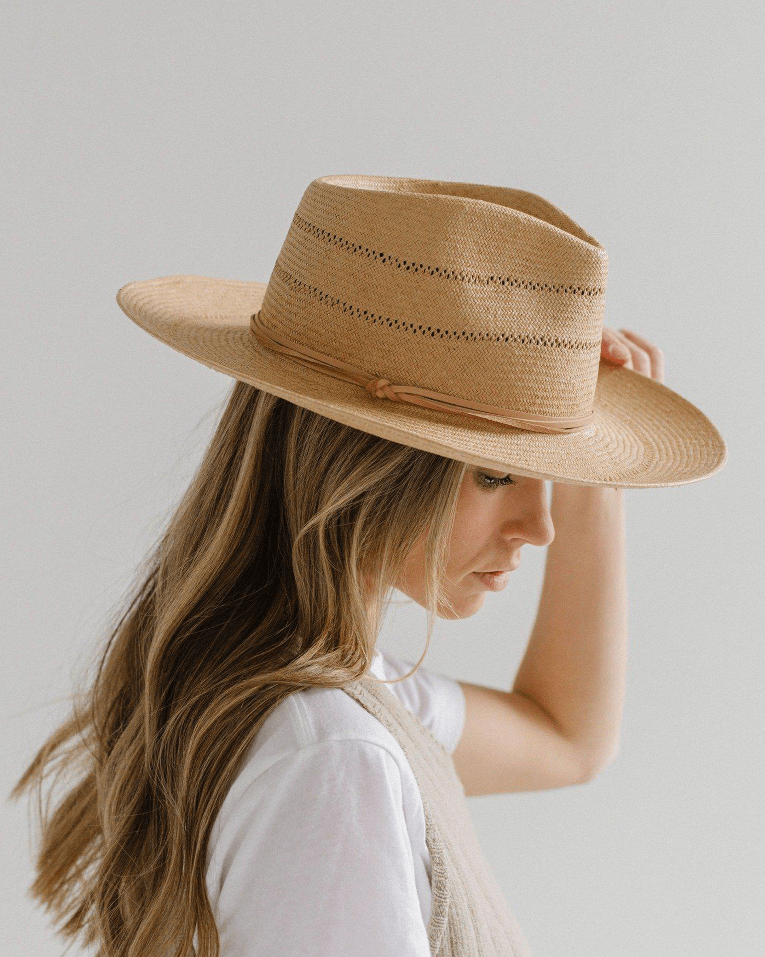 A woman with long, wavy light brown hair wears the Gigi Pip Arlo Honey Straw Teardrop Fedora with a nude band—handwoven venting and adjustable strap visible—as she gazes down, gently touching the brim against a plain background.