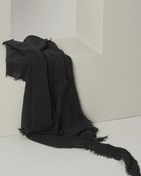The Grisal Love Cashmere Scarf in Black X Charcoal, featuring fringed trims and a soft texture, is draped elegantly over a white geometric rectangular structure against a neutral background.