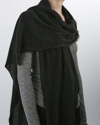 A person wears the Grisal Love Cashmere Scarf in Black X Charcoal, featuring fringed trims, draped over a light gray long-sleeve shirt; shown from shoulders to waist against a plain background.