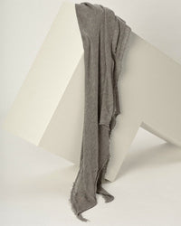 The Grisal Love Cashmere Scarf in Greige, an heirloom-quality accessory with frayed edges, is draped over a white geometric block against a plain background.