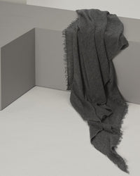 The Grisal Love Cashmere Scarf in Jaspe, a dark gray, heirloom-quality piece, drapes elegantly over the corner of a light gray modern bench, its luxurious length cascading gently to the floor.