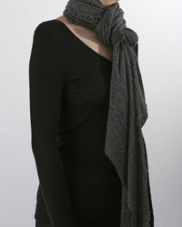 A person wearing a long-sleeved black top and the Grisal Love Cashmere Scarf in Jaspe, loosely wrapped, stands against a plain light background. The person's head is not visible.