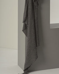 The Grisal Love Cashmere Scarf in Pepper, a dark gray luxury knit with frayed edges, drapes over a modern gray geometric structure against a minimal light background.