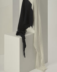 The Grisal Love Duo Cashmere Scarf in Black & Milk, featuring frayed edges, is draped over a white geometric block, creating a bold contrast against the minimal, light-toned background.
